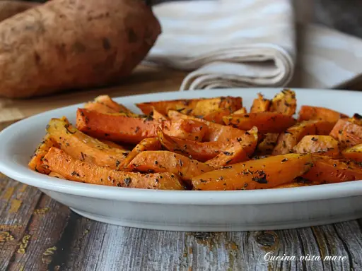 Patate dolci in friggitrice ad aria, tagliate a spicchi e condite con erbe aromatiche, servite in un piatto bianco.