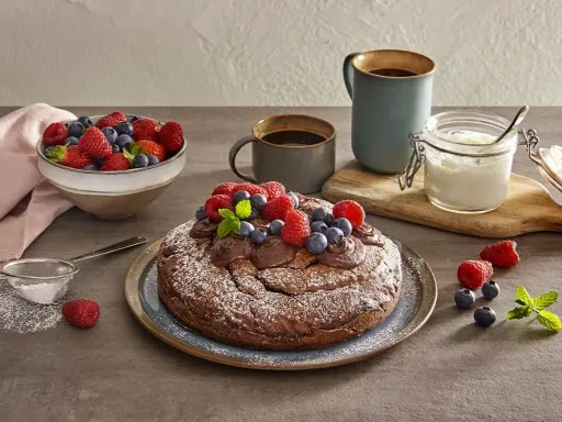 Torta yogurt e cioccolato, cosparsa di zucchero a velo e decorata con frutti di bosco freschi, pronta per essere gustata con un caffè.