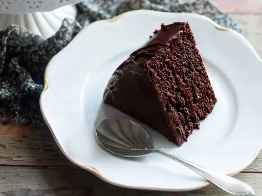 Una fetta di torta al cioccolato morbida e umida, ricoperta di glassa lucida, su un piatto bianco con un cucchiaino.