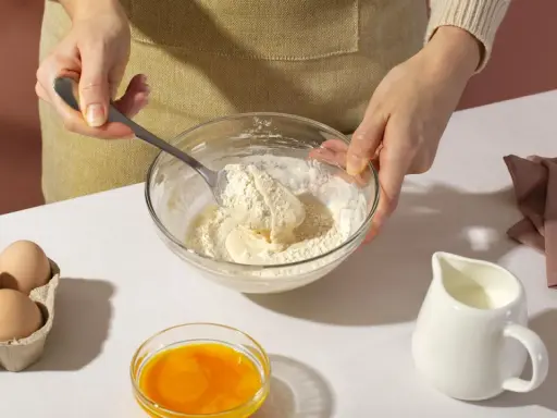 Mani che mescolano farina e burro in una ciotola, con uova, latte e bicarbonato al posto del lievito pronti per la preparazione.