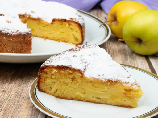 Una fetta di torta di mele invisibile, soffice e profumata, cosparsa di zucchero a velo. Accanto, mele fresche e un tovagliolo viola.