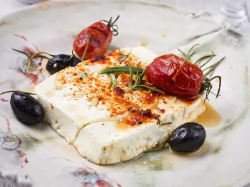 Feta al forno con pomodorini arrostiti, olive nere e rosmarino su un piatto rustico.