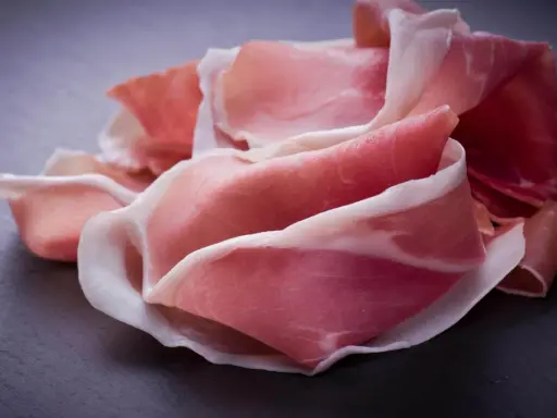 Fette di prosciutto crudo, un insaccato dal colore rosa intenso e venature bianche, disposte su una superficie scura.