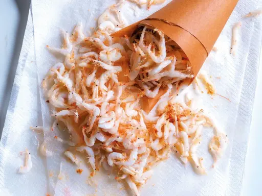 Piccolo cono di carta rovesciato su carta assorbente, con tanti gamberetti surgelati come cucinarli sparsi.