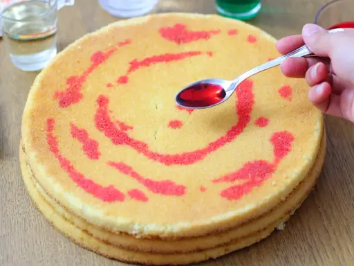 Un cucchiaino di sciroppo rosso viene versato su un pan di spagna decorato con un motivo a spirale. La bagna per pan di spagna renderà la torta più umida e saporita.