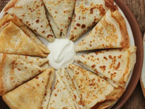 Piatto di crespelle ripiene, piegate a spicchi, con una generosa porzione di panna acida al centro.