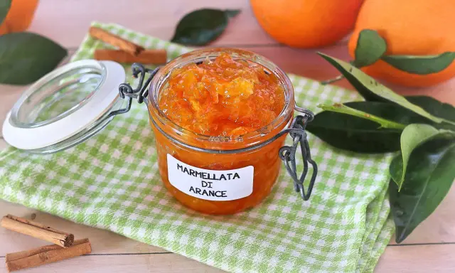 Barattolo di marmellata di arance fatto in casa, con arance fresche e bastoncini di cannella sullo sfondo.