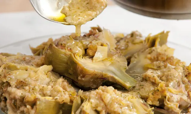 Un cucchiaio versa un sughetto dorato su un piatto di carciofi alla romana, un classico della cucina romana.