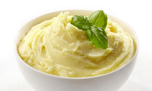 Una ciotola bianca piena di soffice crema di patate, guarnita con foglie di basilico fresco.