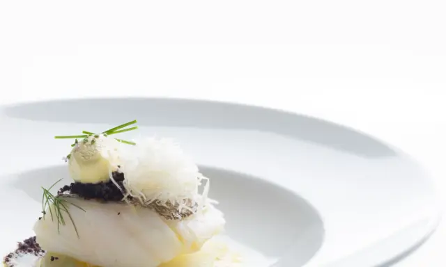Piatto gourmet con filetto di pesce bianco, crema gialla, erbe aromatiche e fili croccanti. Un'interpretazione moderna che evoca la differenza tra baccalà e stoccafisso.