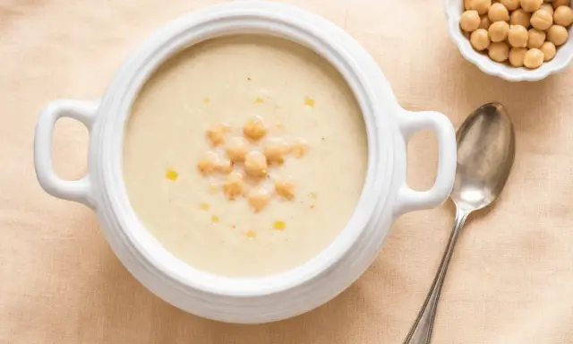 Zuppa cremosa di riso e ceci, guarnita con ceci interi e un filo d'olio. Accanto, una ciotolina di ceci e un cucchiaio d'argento.