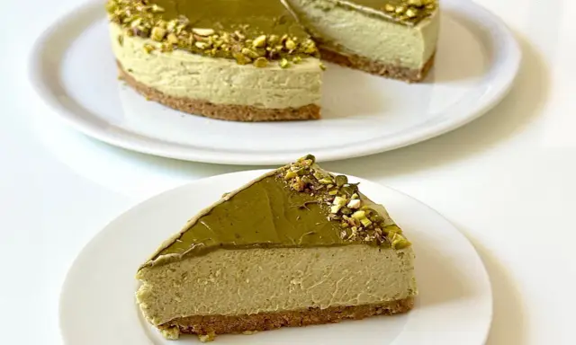 Una fetta di goloso cheesecake al pistacchio, con la torta intera sullo sfondo, decorata con granella di pistacchi.