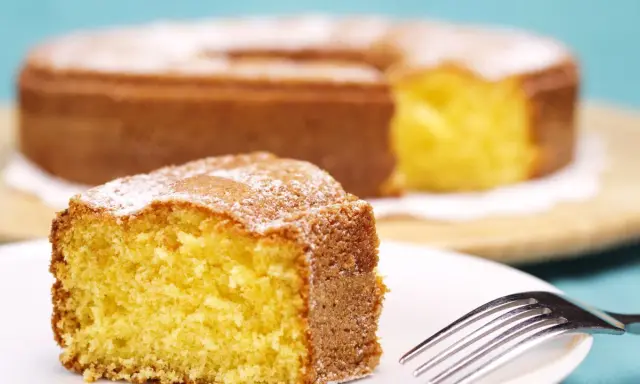 Una fetta di torta 7 vasetti soffice e dorata, cosparsa di zucchero a velo, su un piatto bianco con una forchetta.