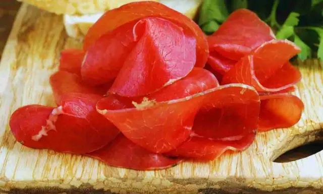 Fette sottili di bresaola, un insaccato di manzo stagionato, disposte su un tagliere di legno con pane e prezzemolo sullo sfondo.