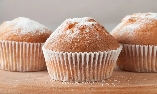 Tre deliziosi muffin senza glutine, spolverati di zucchero a velo, su un tagliere di legno.