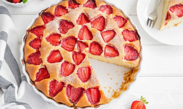 Torta alle fragole dorata, cosparsa di zucchero a velo, con fragole fresche affondate nell'impasto soffice. Una fetta è già stata servita.