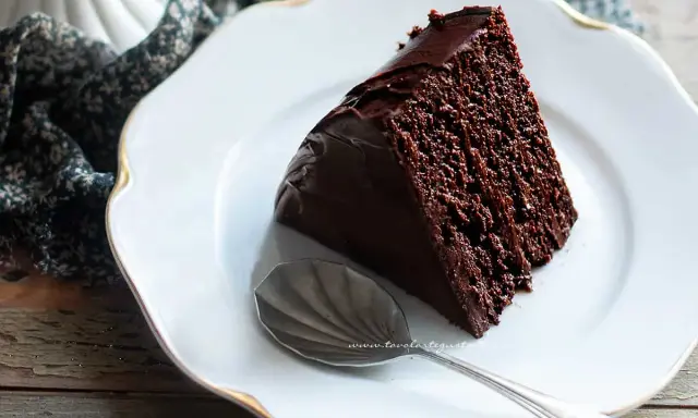 Una fetta di torta al cioccolato morbida e umida, ricoperta di glassa lucida, su un piatto bianco con un cucchiaino.