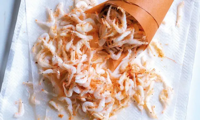 Piccolo cono di carta rovesciato su carta assorbente, con tanti gamberetti surgelati come cucinarli sparsi.