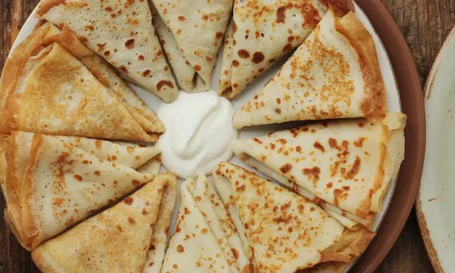 Piatto di crespelle ripiene, piegate a spicchi, con una generosa porzione di panna acida al centro.