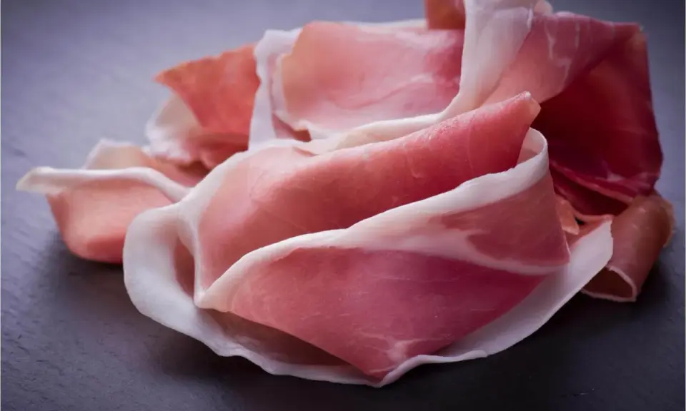 Fette di prosciutto crudo, un insaccato dal colore rosa intenso e venature bianche, disposte su una superficie scura.