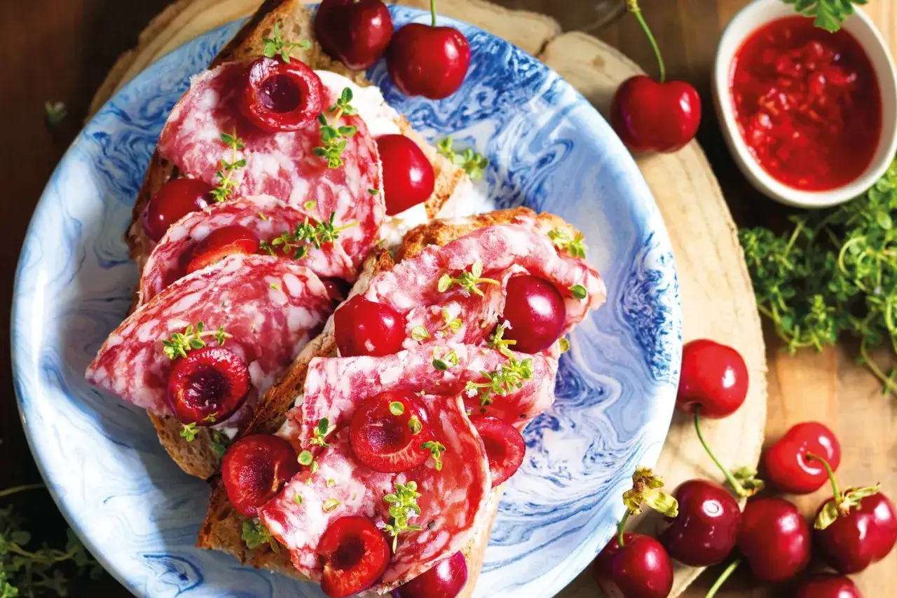 Bruschette con salame e variet&agrave; ciliegie giganti, guarnite con erbe aromatiche. Un contrasto dolce-salato invitante.