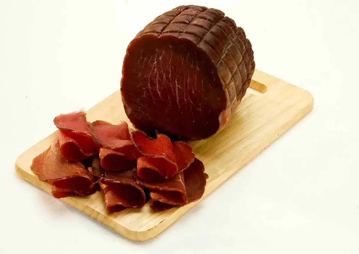 Un pezzo intero e fette arrotolate di bresaola, un insaccato di carne rossa, su un tagliere di legno.