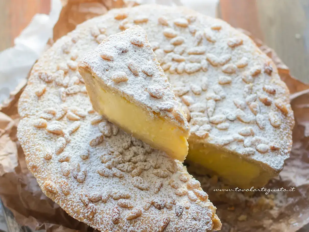 Torta con crema pasticcera, ricoperta di pinoli e zucchero a velo. Una fetta &egrave; stata tagliata, rivelando il ripieno cremoso.