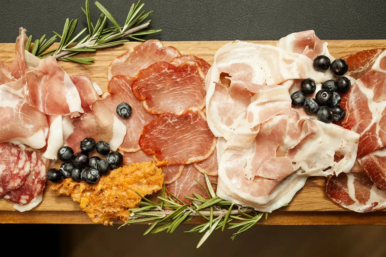 Assortimento di salumi: prosciutto, pancetta, capocollo e altri tipi di salame, guarniti con mirtilli e rosmarino.