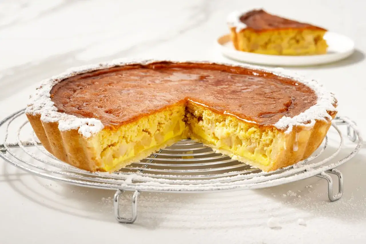 Una fetta di torta di mele dorata, con ripieno cremoso e mele a pezzi, spolverata di zucchero a velo.
