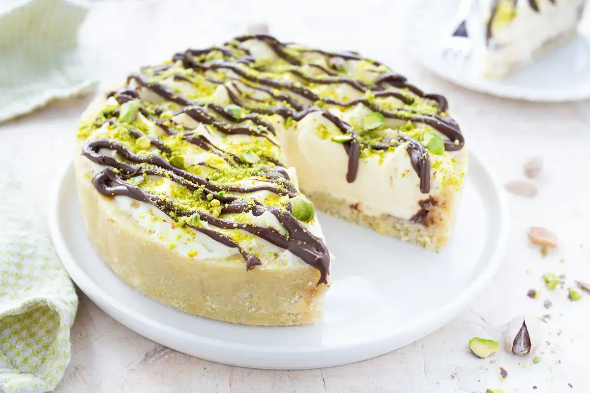Deliziosa cheesecake al pistacchio con glassa al cioccolato e granella verde brillante. Una fetta &egrave; gi&agrave; stata servita, invitando a gustare questo dolce.