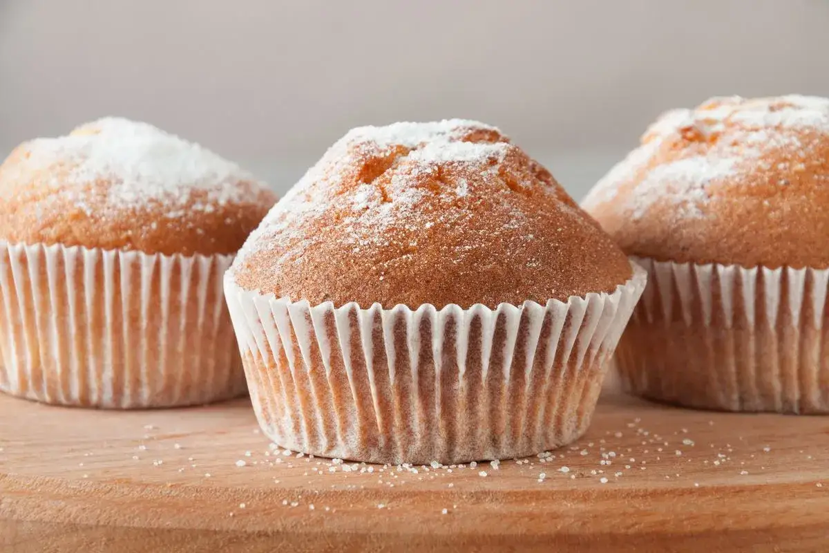 Tre deliziosi muffin senza glutine, spolverati di zucchero a velo, su un tagliere di legno.