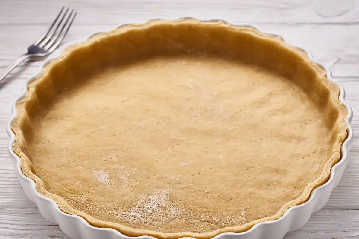 Base per crostata pronta, una deliziosa pasta frolla senza glutine, in una teglia bianca con forchetta sullo sfondo.