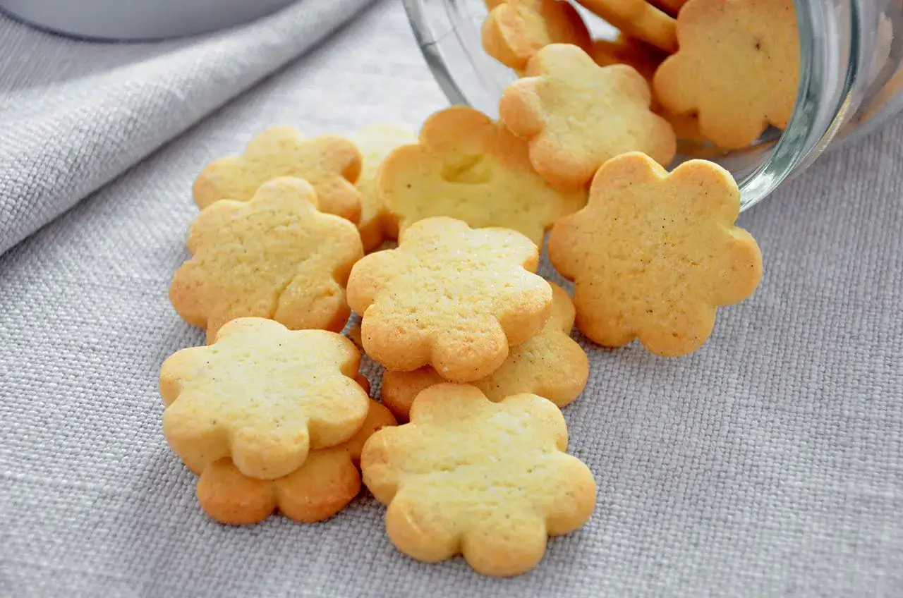 Biscotti al burro a forma di fiore, dorati e fragranti, sparsi su una tovaglia grigia.