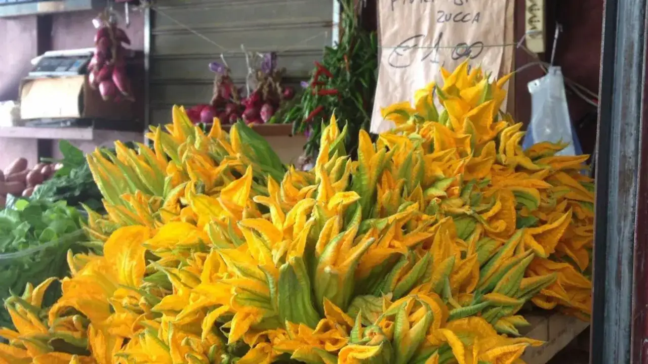 Un cesto traboccante di vivaci fiori di zucca gialli e verdi, pronti per essere cucinati. Sullo sfondo, cipolle rosse e verdure fresche.