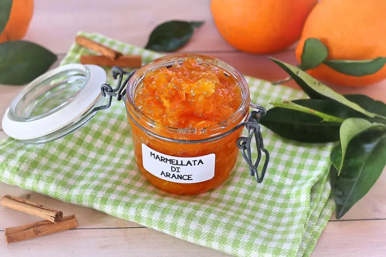Barattolo di marmellata di arance fatto in casa, con arance fresche e bastoncini di cannella sullo sfondo.