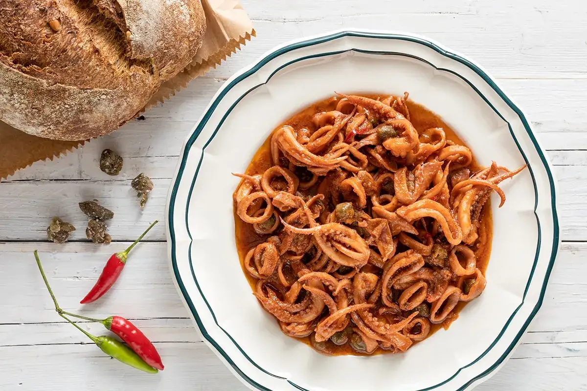 Un piatto invitante di totani in umido, con capperi e peperoncino, accanto a un pane rustico.