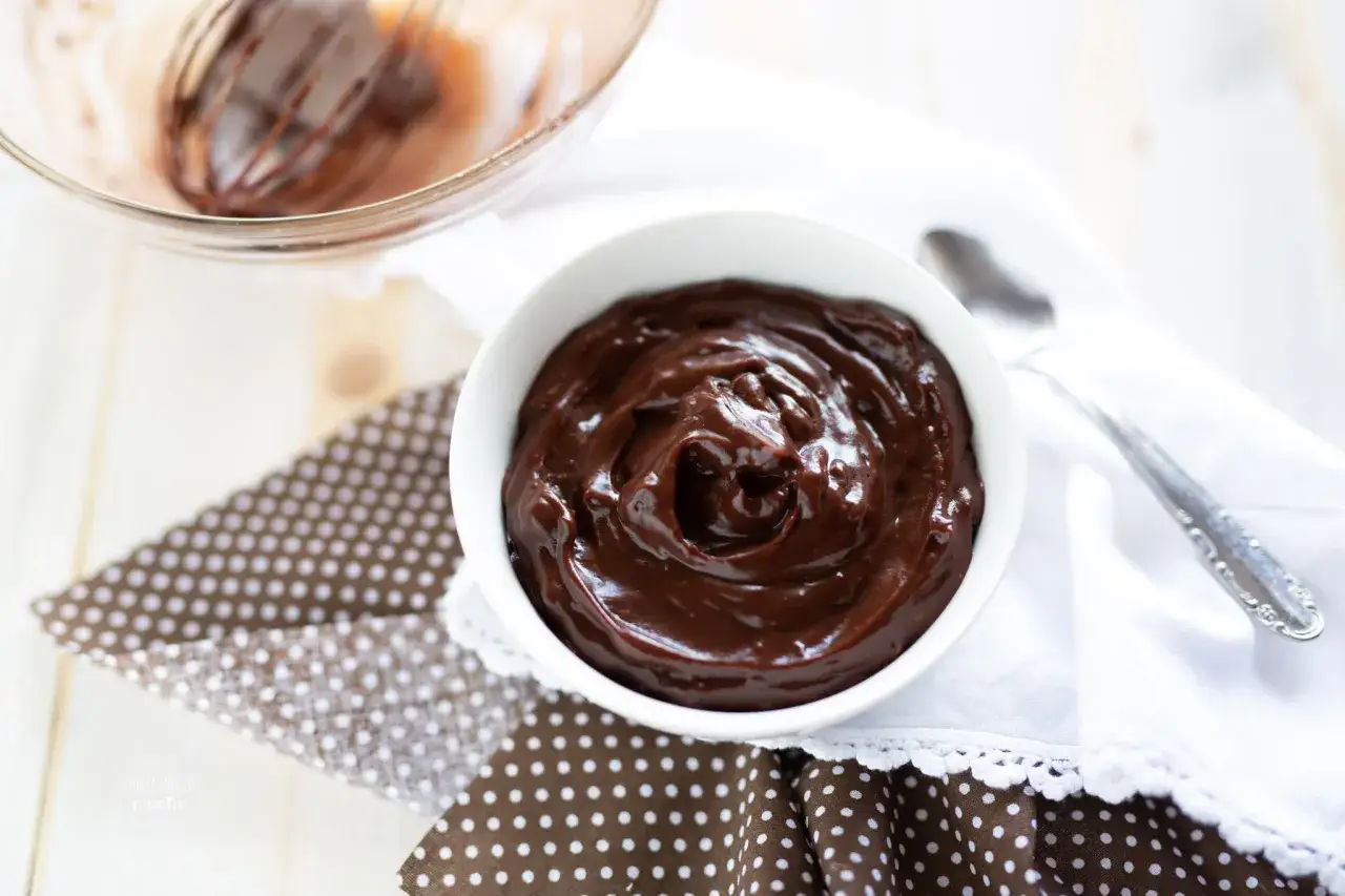 Una ciotola bianca piena di lucida crema al cioccolato, appena preparata con una frusta in una ciotola di vetro sullo sfondo.