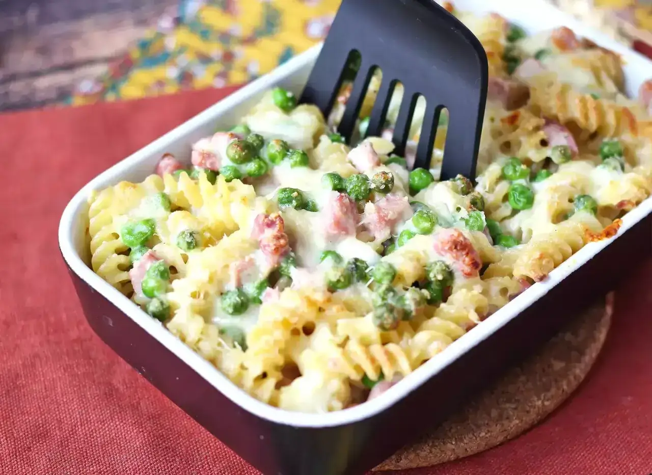 Un piatto invitante di pasta pasticciata al forno con piselli e prosciutto, servita in una pirofila scura.