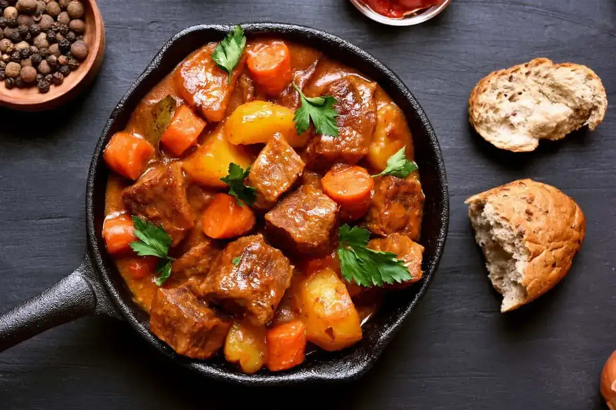 Un saporito capretto in umido con patate e carote, guarnito con prezzemolo fresco, servito in una padella di ghisa. Accanto, pane rustico e una ciotola di pepe in grani.
