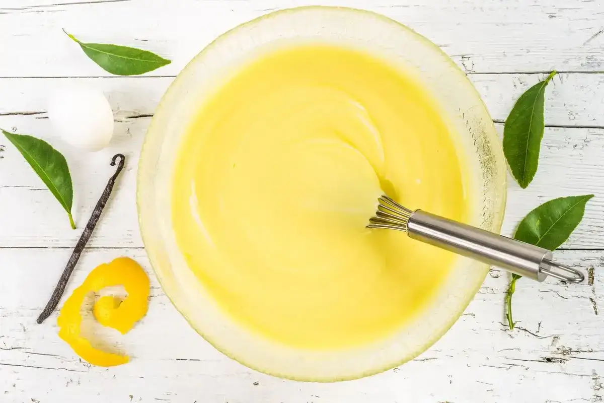 Una ciotola di uovo sbattuto con zucchero, con accanto un uovo, una bacca di vaniglia, scorza di limone e foglie verdi.