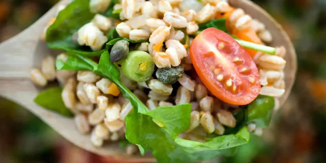 Un cucchiaio di legno pieno di insalata di farro, pomodoro, spinaci e capperi. Un piatto sano e gustoso.