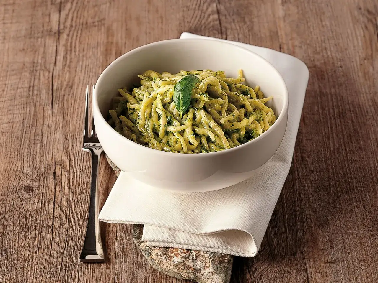 Trenette al pesto, un piatto di pasta fresca condita con un ricco sugo verde, guarnito con una foglia di basilico.