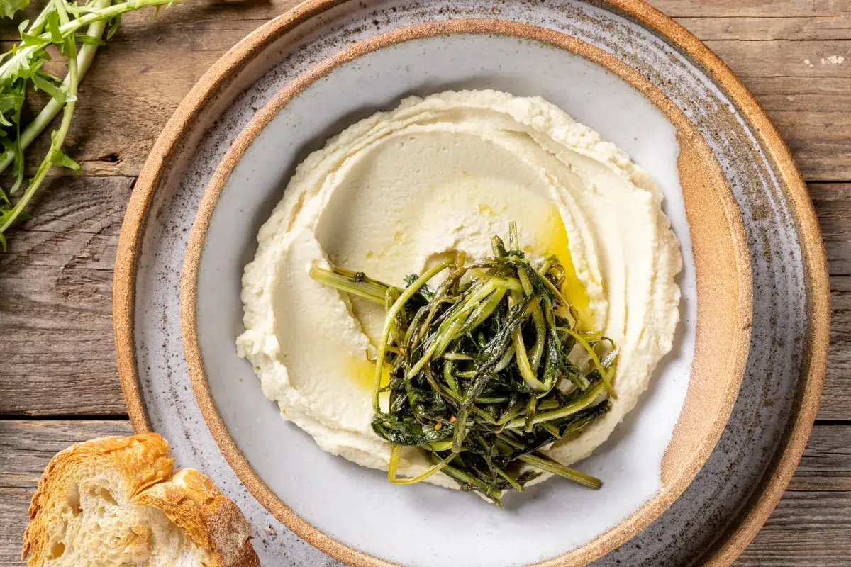 Crema vellutata di pur&egrave; di fave, condita con olio e erbe verdi, servita su un piatto rustico con pane croccante.