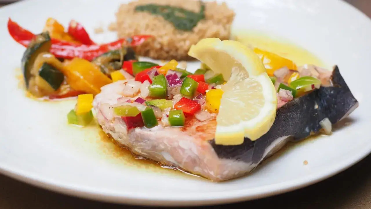 Un succulento pesce spada alla siciliana, guarnito con verdure colorate e una fetta di limone, servito su un piatto bianco.