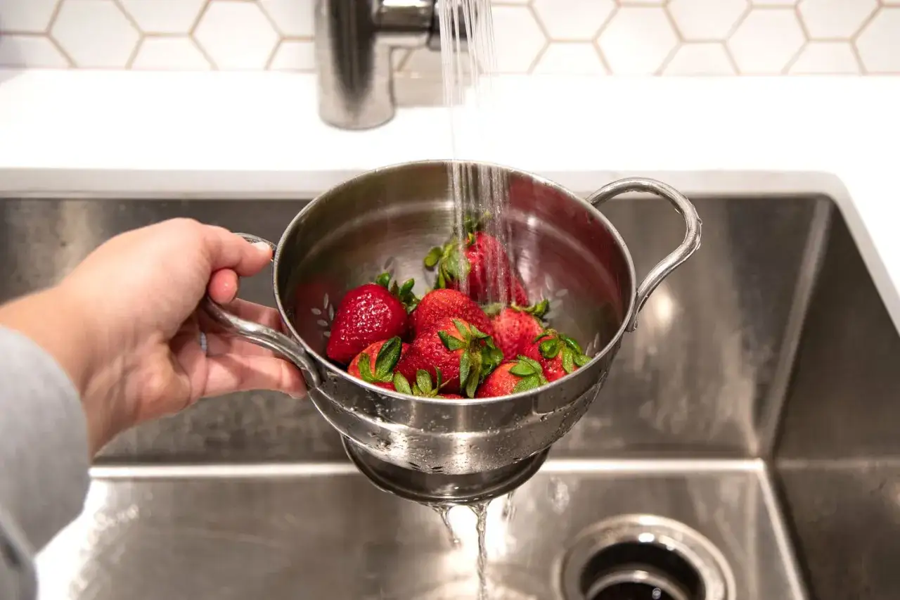 Mano che tiene un colino con fragole rosse sotto un getto d'acqua corrente. Ecco come lavare le fragole per gustarle fresche.