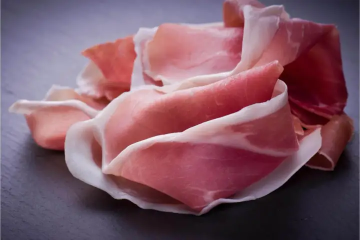 Fette di prosciutto crudo, un insaccato dal colore rosa intenso e venature bianche, disposte su una superficie scura.