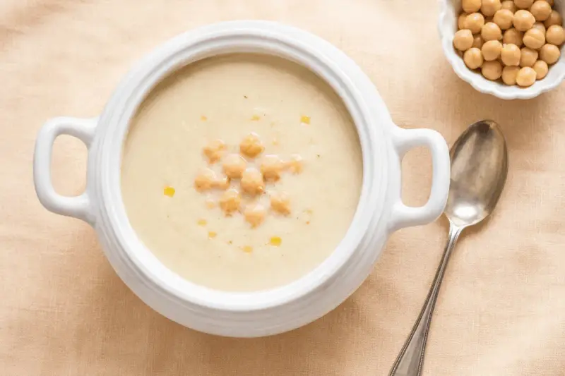 Zuppa cremosa di riso e ceci, guarnita con ceci interi e un filo d'olio. Accanto, una ciotolina di ceci e un cucchiaio d'argento.